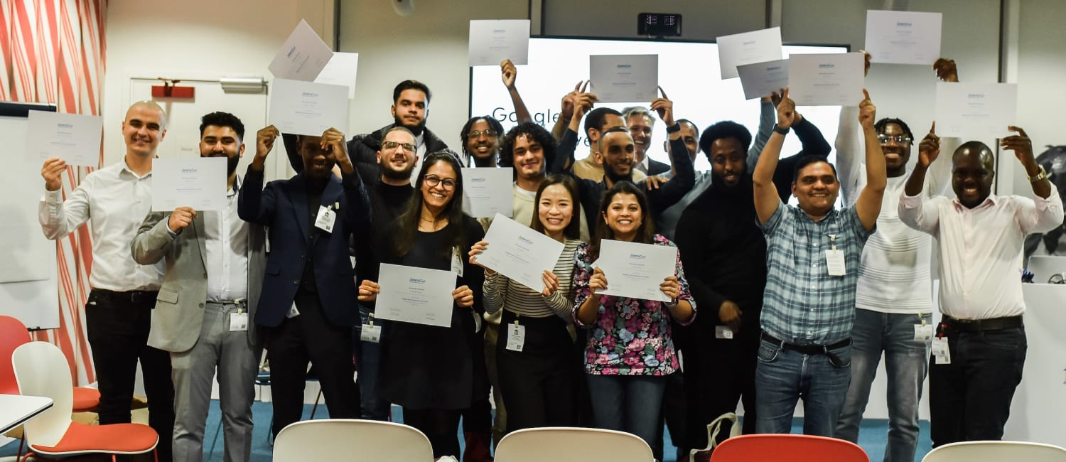 A group of people stand indoors, holding up certificates and smiling for a group photo in front of a presentation screen.